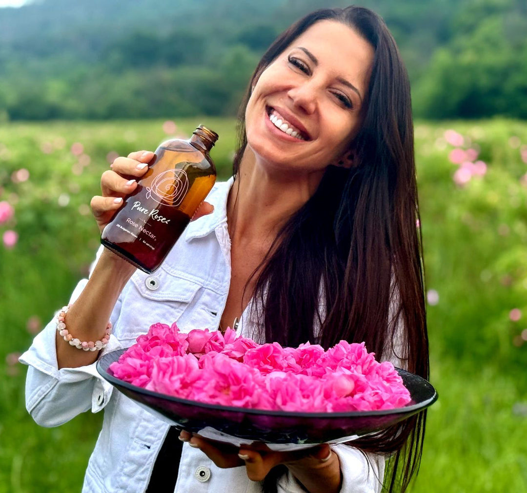 Woman smiling in a Bulgarian rose field, holding a bottle of Rose Nectar and a bowl of pink roses